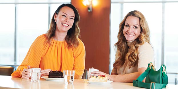 Kaksi naista hymyilee leveästi pöydän ääressä. Heillä on edessään kakkupalat, kupit kahvia ja vesilasilliset.