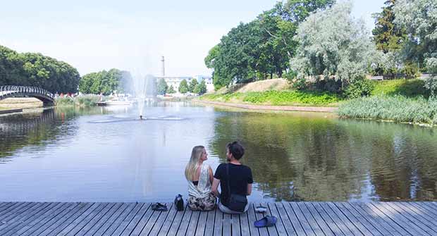 Nuori nainen ja mies istuvat puisella sillalla katsellen lampea. Lammessa on suihkulähde.