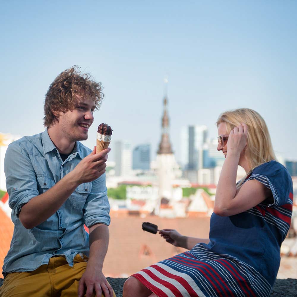 Mies ja nainen istuvat muurilla ja syövät jäätelöä. Taustalla näkyy vanhan kaupungin kirkon torni ja muita rakennuksia.