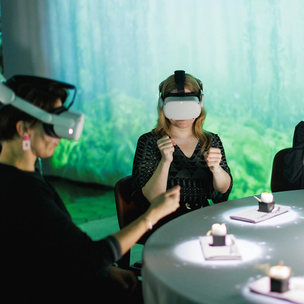 Kaksi naista istuu ruokapöydässä virtuaalitodellisuuslasit päässä.
