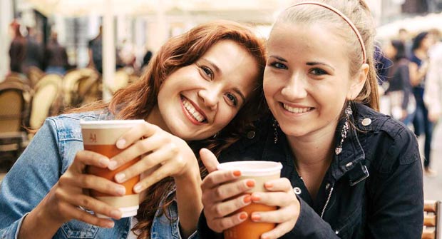 Puna- ja vaaleatukkaiset nuoret naiset hymyilevät kameralle take away -kahvimuki kädessä.