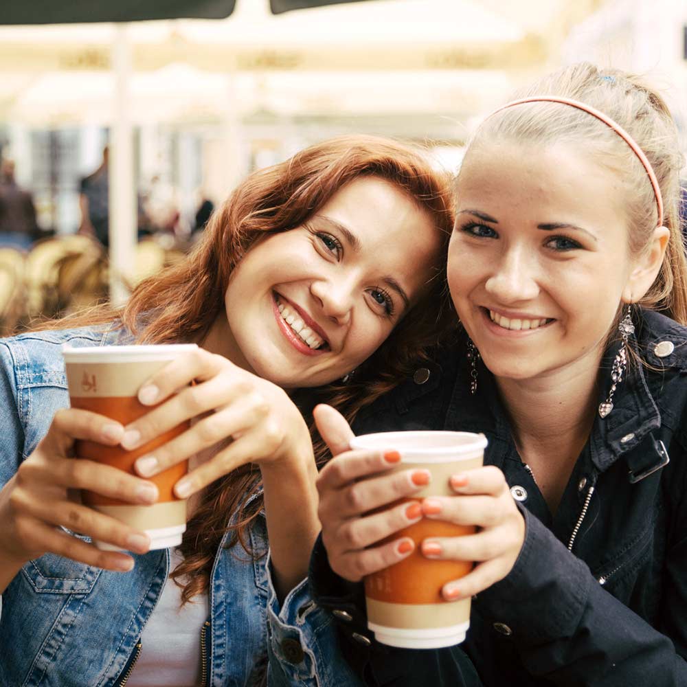 Puna- ja vaaleatukkaiset nuoret naiset hymyilevät kameralle take away -kahvimuki kädessä.